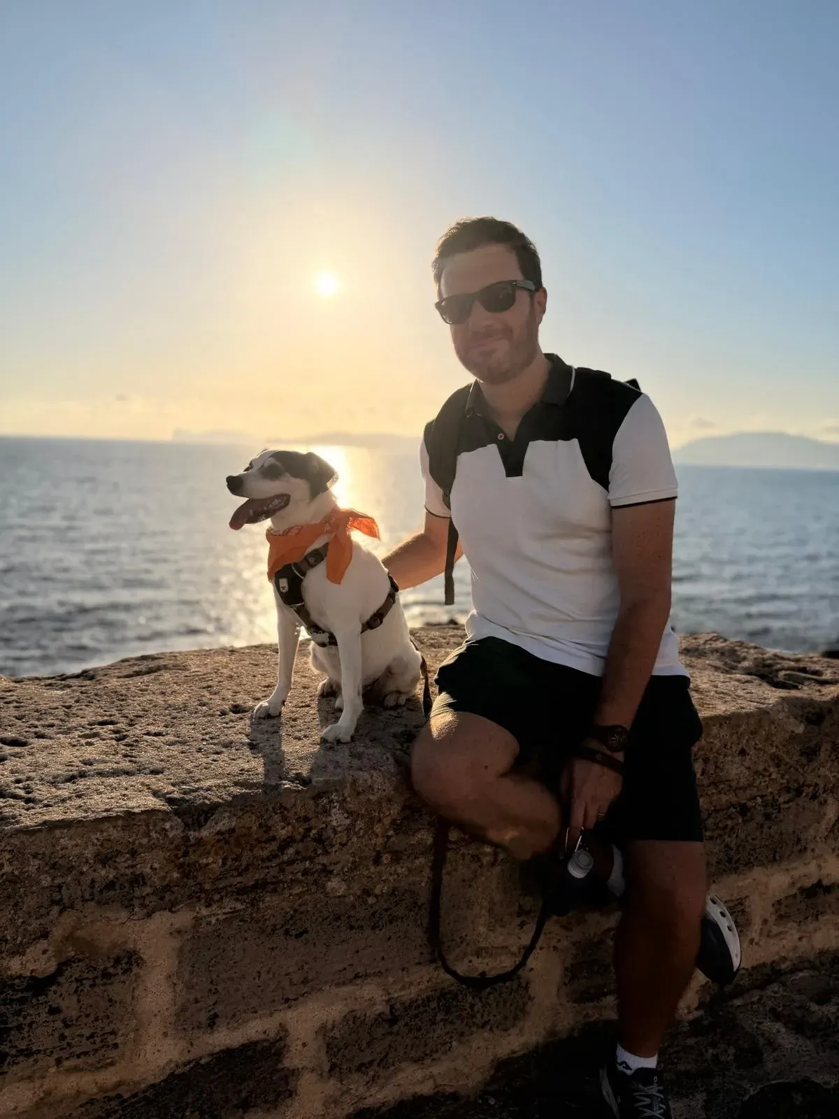 Figaro, un perro blanco y negro con un pañuelo naranja, sentado en un muro de piedra junto al mar al atardecer con Rubén Alejos Dasilva